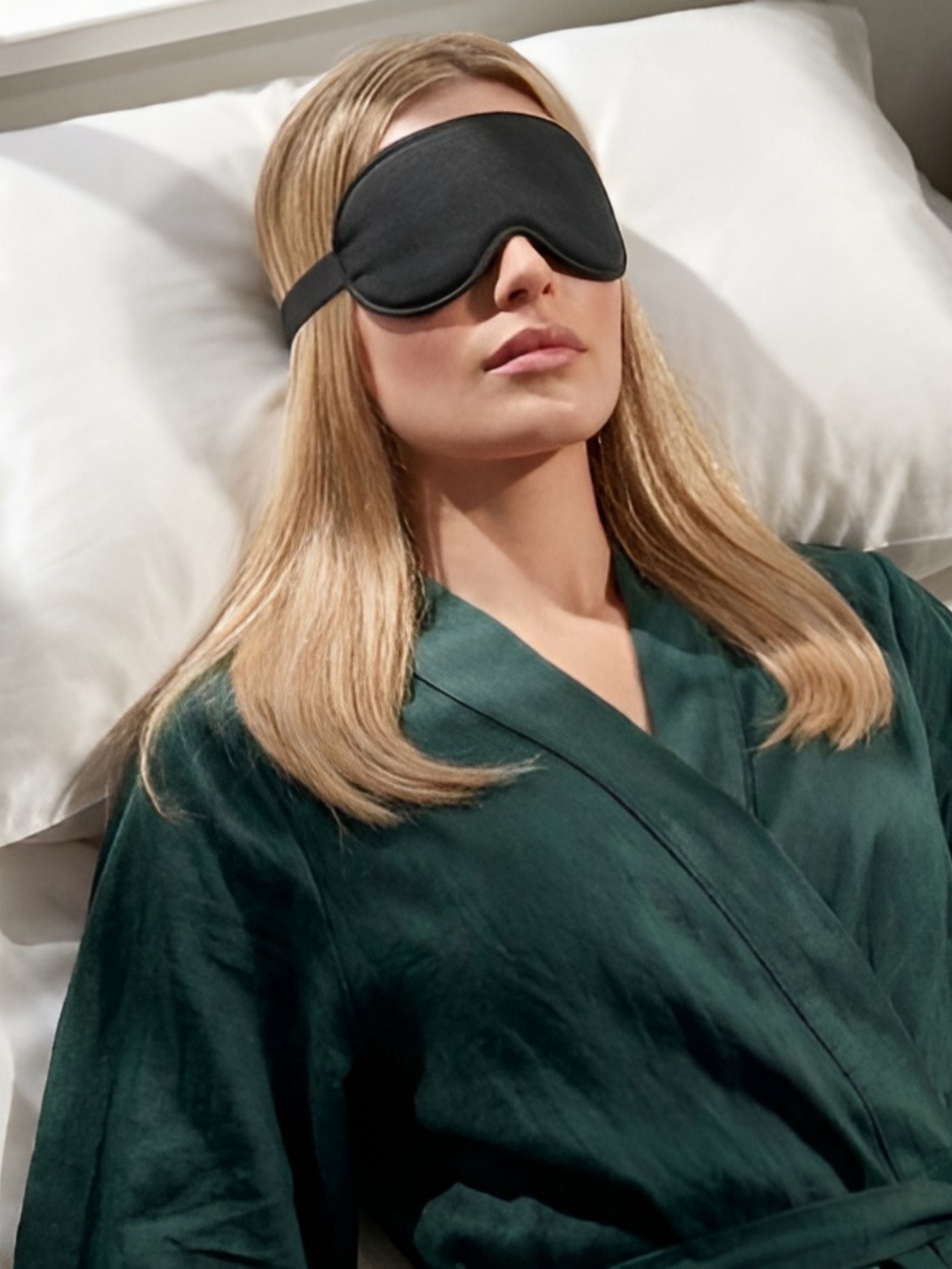Woman wearing a black sleep mask and green robe lying on a white pillow